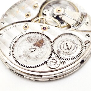 May include: Close-up of a vintage watch movement with intricate gears, screws, and jewels. The silver-toned mechanism features the text "17 Jewels" and "Six Adjustments". The watch components show signs of age and wear.