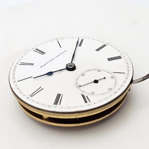 May include: A vintage pocket watch face with a white dial, black Roman numerals, and a small seconds dial. The watch hands are black and blue. The watch casing is gold-toned. The text "E. HOWARD WATCH CO." is visible on the dial.