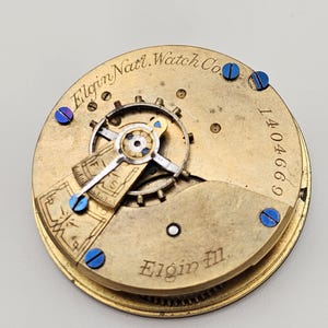 May include: Close-up of a vintage Elgin National Watch Co. movement. The gold-toned mechanism features intricate gears, blue screws, and the engraved text "Elgin Natl. Watch Co." and "Elgin Ill.". The serial number 1404669 is also visible.