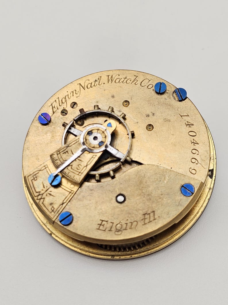 May include: Close-up of a vintage Elgin National Watch Co. movement. The gold-toned mechanism features intricate gears, blue screws, and the engraved text "Elgin Natl. Watch Co." and "Elgin Ill.". The serial number 1404669 is also visible.