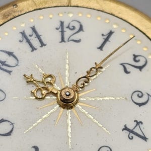 May include: Close-up of a vintage watch face. The white face features black numerals, gold hands, and a gold-colored sunburst design emanating from the center. The watch hands point to approximately 1:50. The watch has a gold-colored bezel.