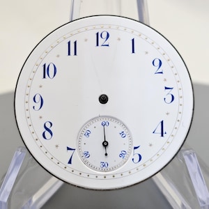 Puede incluir: Esfera de reloj de bolsillo blanca con marcadores de hora en n&uacute;meros romanos azules y agujas negras. El reloj tiene un peque&ntilde;o subdial blanco con marcas negras para los segundos.