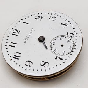 May include: A vintage Elgin pocket watch face with a white background, black numerals, and a smaller seconds dial. The watch face has a metal frame and a small hand. The word "Elgin" is printed on the face.