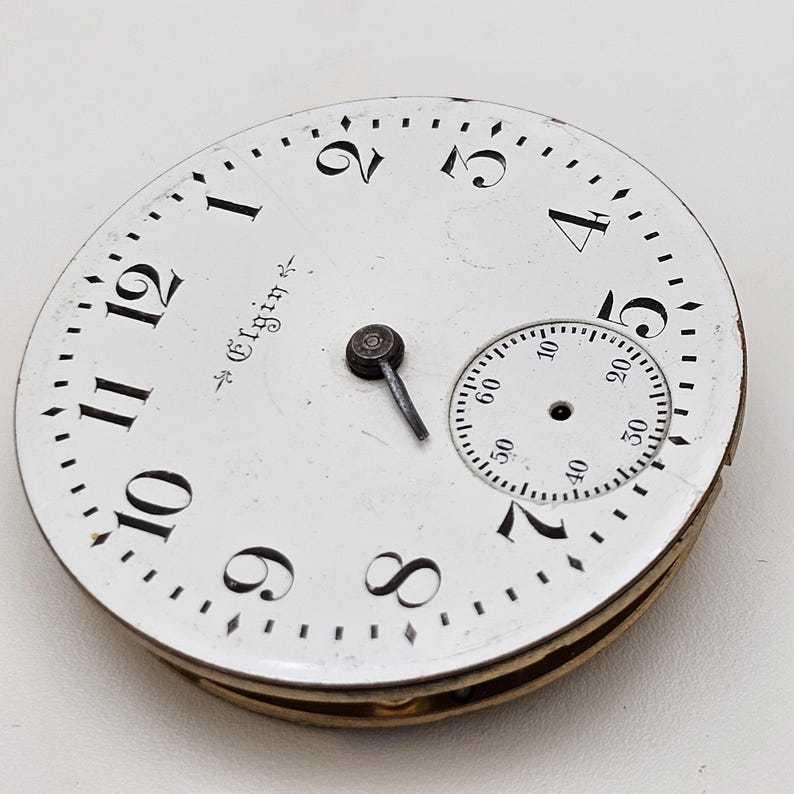 May include: A vintage Elgin pocket watch face with a white background, black numerals, and a smaller seconds dial. The watch face has a metal frame and a small hand. The word "Elgin" is printed on the face.