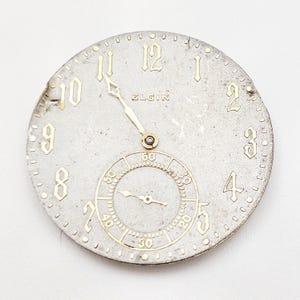 May include: A vintage Elgin watch face with a silver-tone finish and gold-colored numerals and hands. The face includes a smaller subdial for seconds and the brand name "ELGIN". The watch face is round and shows signs of age.