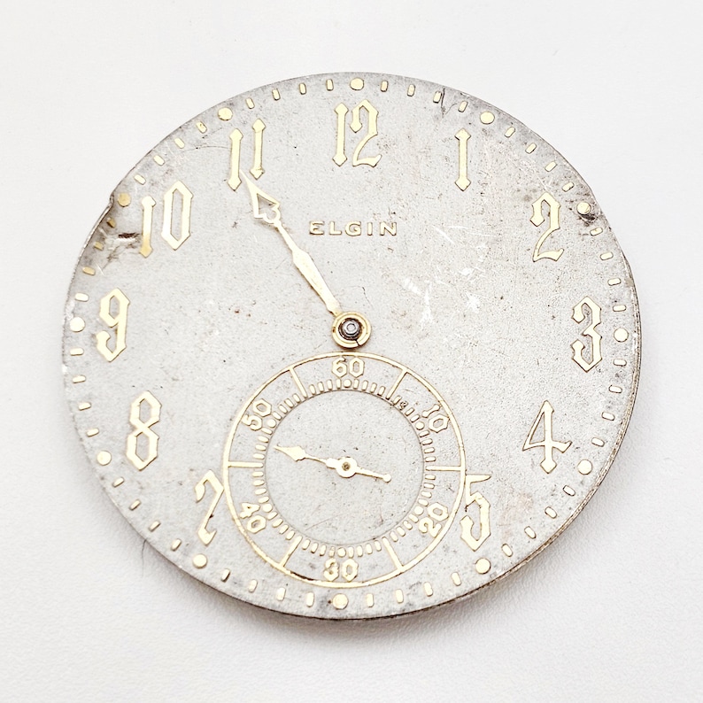 May include: A vintage Elgin watch face with a silver-tone finish and gold-colored numerals and hands. The face includes a smaller subdial for seconds and the brand name "ELGIN". The watch face is round and shows signs of age.