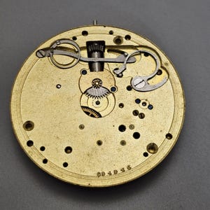 May include: Close-up of a gold-colored watch movement with intricate gears and levers. The movement is visible through a clear glass cover. The watch movement is labeled with the number "694945".