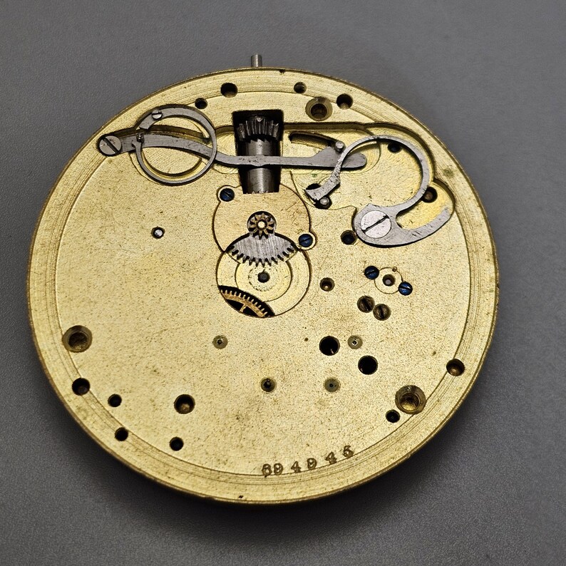 May include: Close-up of a gold-colored watch movement with intricate gears and levers. The movement is visible through a clear glass cover. The watch movement is labeled with the number "694945".