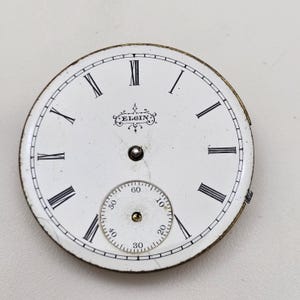 以下が含まれることがあります： 白い背景に黒いローマ数字と小さな秒針が付いた、アンティークのエルジン時計の文字盤。文字盤には装飾的なフォントで「ELGIN」の文字が入っています。文字盤は丸く、金色の金属製の縁があります。