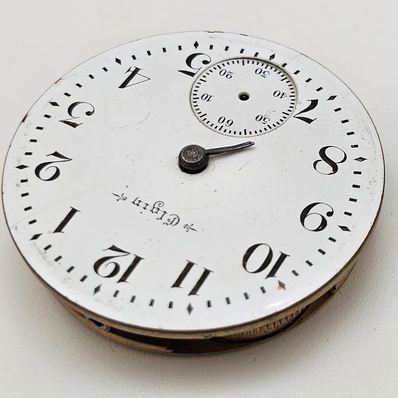 May include: Close-up of an antique Elgin pocket watch face. The white face features black numerals, a smaller seconds dial, and a black hand. The word "Elgin" is printed on the face.