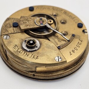May include: Close-up of an antique Elgin III pocket watch movement. The gold-toned mechanism features intricate details, including gears, screws, and the text "Elgin Ill." and "Elgin Natl. Watch Co." The serial number "725547" is visible.