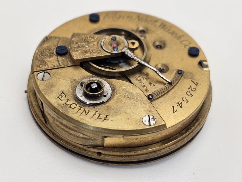 May include: Close-up of an antique Elgin III pocket watch movement. The gold-toned mechanism features intricate details, including gears, screws, and the text "Elgin Ill." and "Elgin Natl. Watch Co." The serial number "725547" is visible.