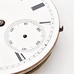 Puede incluir: Primer plano de una esfera de reloj vintage con fondo blanco. La esfera presenta n&uacute;meros romanos negros, indicadores de minutos y una esfera m&aacute;s peque&ntilde;a con n&uacute;meros. El reloj tiene un borde met&aacute;lico y un peque&ntilde;o componente met&aacute;lico.