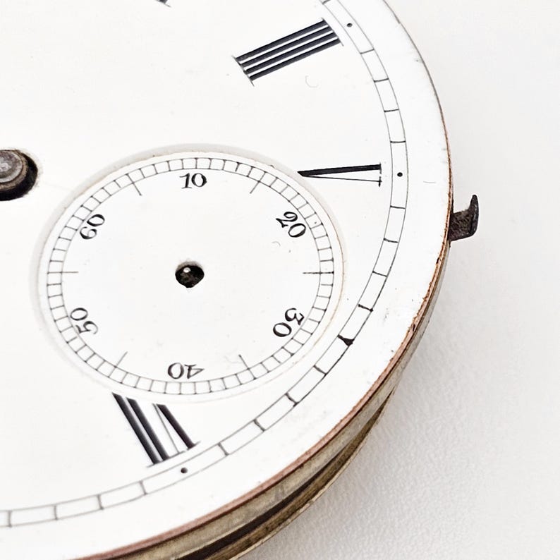 Puede incluir: Primer plano de una esfera de reloj vintage con fondo blanco. La esfera presenta n&uacute;meros romanos negros, indicadores de minutos y una esfera m&aacute;s peque&ntilde;a con n&uacute;meros. El reloj tiene un borde met&aacute;lico y un peque&ntilde;o componente met&aacute;lico.