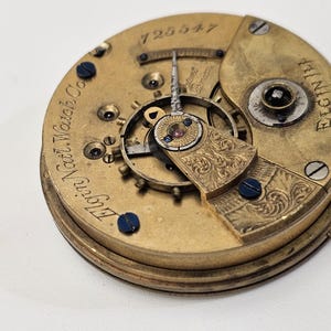 May include: Close-up of a vintage watch movement. The gold-toned mechanism features intricate gears, screws, and engraved details. The text "Elgin Natl. Watch Co." and "Elgin Ill" are visible, along with the number "725547".