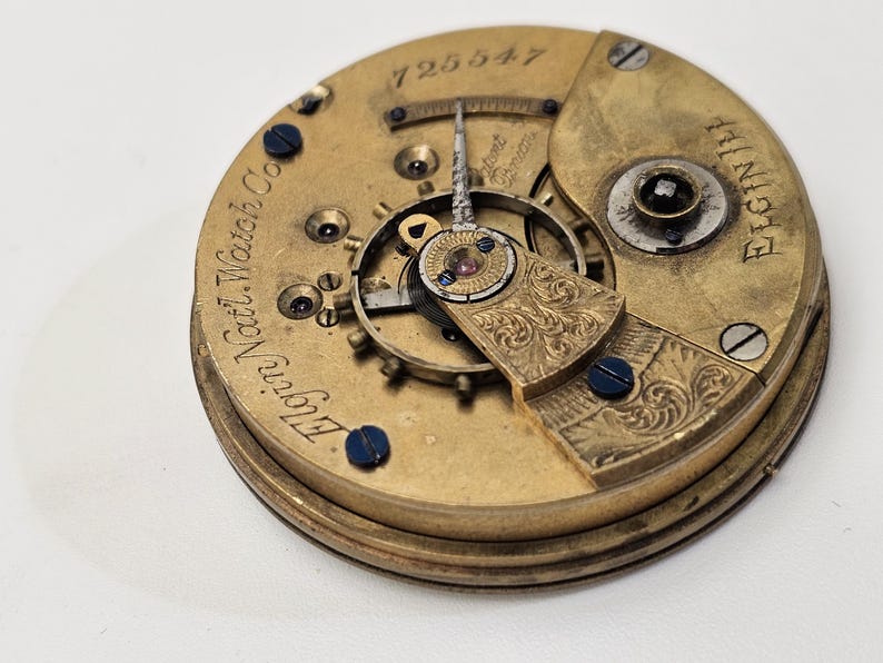 May include: Close-up of a vintage watch movement. The gold-toned mechanism features intricate gears, screws, and engraved details. The text "Elgin Natl. Watch Co." and "Elgin Ill" are visible, along with the number "725547".