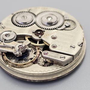 Puede incluir: Vista cercana de un movimiento de reloj de bolsillo plateado con engranajes intrincados y un mecanismo de cuerda. La esfera del reloj no es visible en esta imagen.