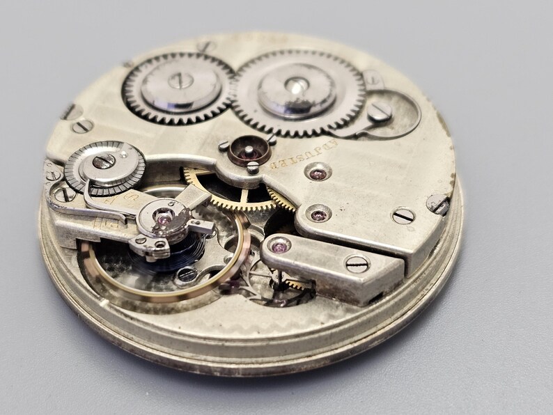 Puede incluir: Vista cercana de un movimiento de reloj de bolsillo plateado con engranajes intrincados y un mecanismo de cuerda. La esfera del reloj no es visible en esta imagen.