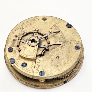 Puede incluir: Primer plano de un movimiento de reloj antiguo. El mecanismo de lat&oacute;n presenta el texto "SPRINGFIELD, MASS" y "PATENT PINION". Tambi&eacute;n es visible el n&uacute;mero de serie "329437". El movimiento del reloj tiene engranajes intrincados y tornillos azules.