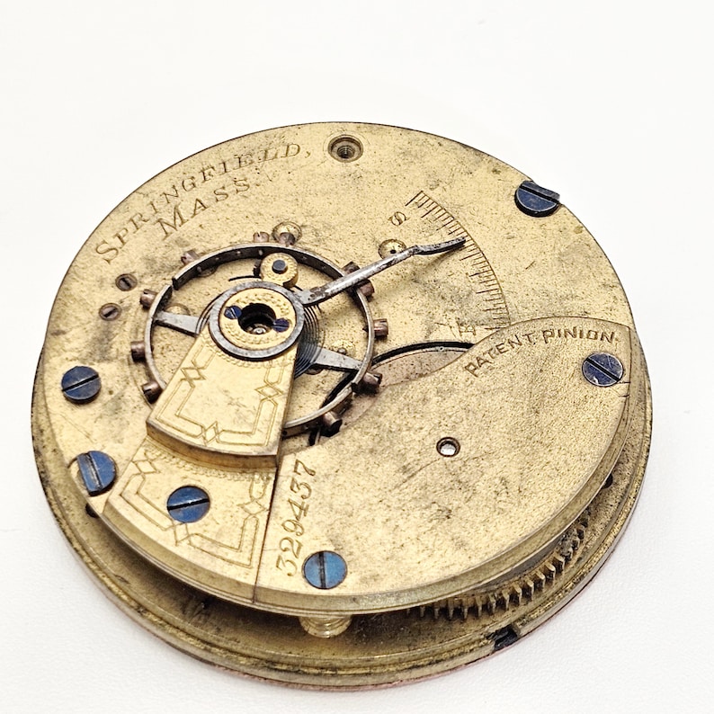 Puede incluir: Primer plano de un movimiento de reloj antiguo. El mecanismo de lat&oacute;n presenta el texto "SPRINGFIELD, MASS" y "PATENT PINION". Tambi&eacute;n es visible el n&uacute;mero de serie "329437". El movimiento del reloj tiene engranajes intrincados y tornillos azules.