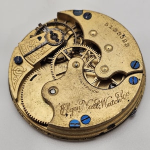 以下が含まれることがあります： ヴィンテージのElgin Natl. Watch Co.の時計ムーブメントのクローズアップ。金色のメカニズムは、複雑な歯車、ネジ、シリアル番号を備えています。青いネジが円形のデザインを強調しています。精密工学の証です。