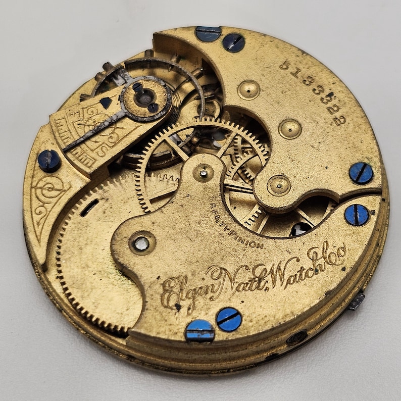 以下が含まれることがあります： ヴィンテージのElgin Natl. Watch Co.の時計ムーブメントのクローズアップ。金色のメカニズムは、複雑な歯車、ネジ、シリアル番号を備えています。青いネジが円形のデザインを強調しています。精密工学の証です。