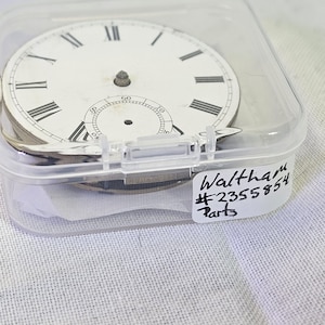 Puede incluir: Una esfera de reloj blanca con marcadores de hora en n&uacute;meros romanos negros y marcadores de minutos negros. La esfera del reloj est&aacute; en un contenedor de pl&aacute;stico transparente con una etiqueta blanca que dice "Waltham #2355854 Parts".