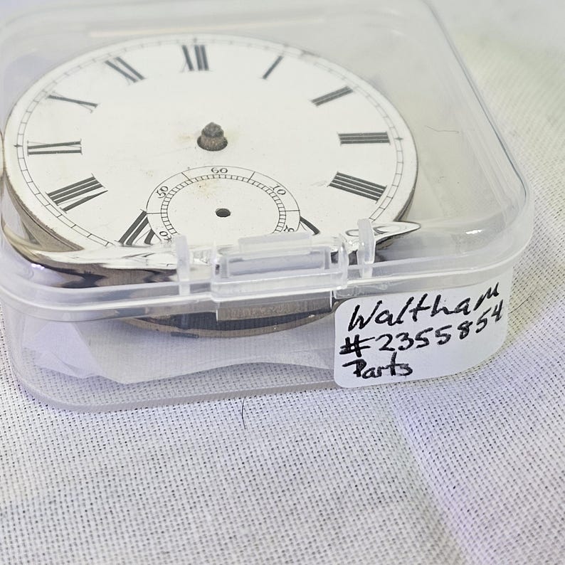 Puede incluir: Una esfera de reloj blanca con marcadores de hora en n&uacute;meros romanos negros y marcadores de minutos negros. La esfera del reloj est&aacute; en un contenedor de pl&aacute;stico transparente con una etiqueta blanca que dice "Waltham #2355854 Parts".