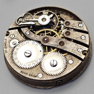 May include: Close-up of a vintage watch movement. The mechanism features numerous gears, screws, and a balance wheel. The watch face is marked with "Tavannes Watch Co" and "Swiss Made". The movement has 17 jewels.