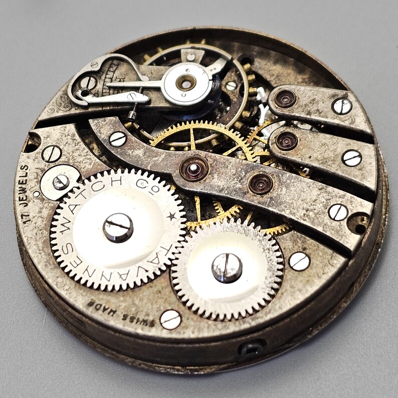 May include: Close-up of a vintage watch movement. The mechanism features numerous gears, screws, and a balance wheel. The watch face is marked with "Tavannes Watch Co" and "Swiss Made". The movement has 17 jewels.