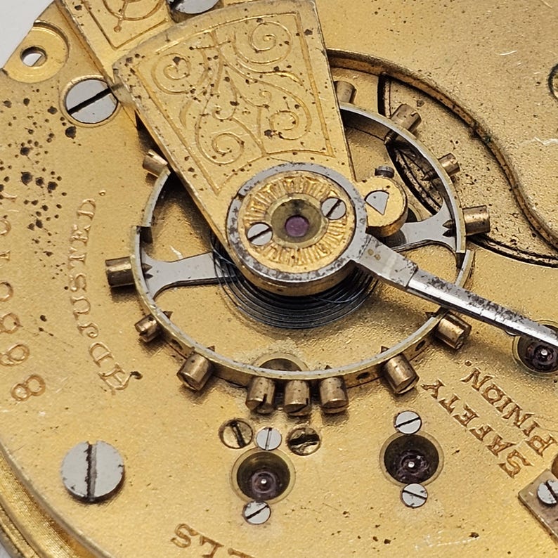 May include: Close-up of a vintage watch movement with intricate details. The gold-toned mechanism features gears, screws, and a balance wheel. The words "Adjusted" and "Safety Pinion" are visible on the watch face. The watch has a classic, antique aesthetic.