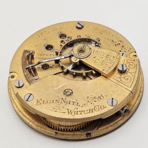May include: Close-up of a vintage Elgin National Watch Co. watch movement. The gold-toned mechanism features intricate gears, screws, and the text "17 Jewels" and "Safety Pinion". The watch movement is a detailed example of early 20th-century watchmaking.