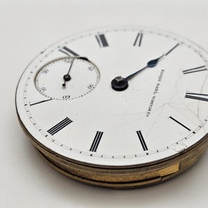 May include: Close-up of an antique pocket watch face. The white face features black Roman numerals, a smaller seconds dial, and the text "Elgin Natl. Watch Co." The watch has a gold-colored metal casing and black hands.
