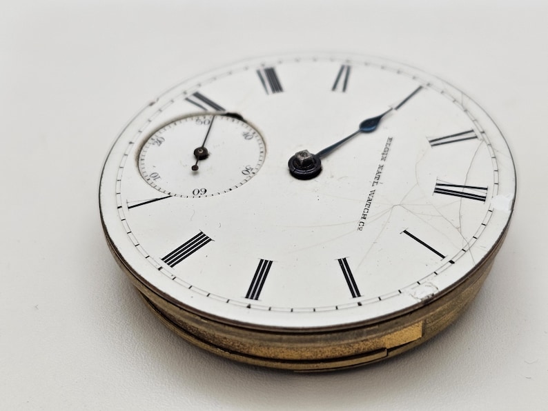 May include: Close-up of an antique pocket watch face. The white face features black Roman numerals, a smaller seconds dial, and the text "Elgin Natl. Watch Co." The watch has a gold-colored metal casing and black hands.