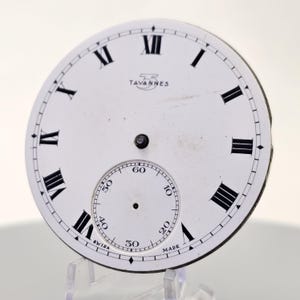 May include: A white watch face with black Roman numerals and a smaller seconds dial. The text "TAVANNES" is printed above the center. The words "SWISS MADE" are printed at the bottom of the seconds dial.