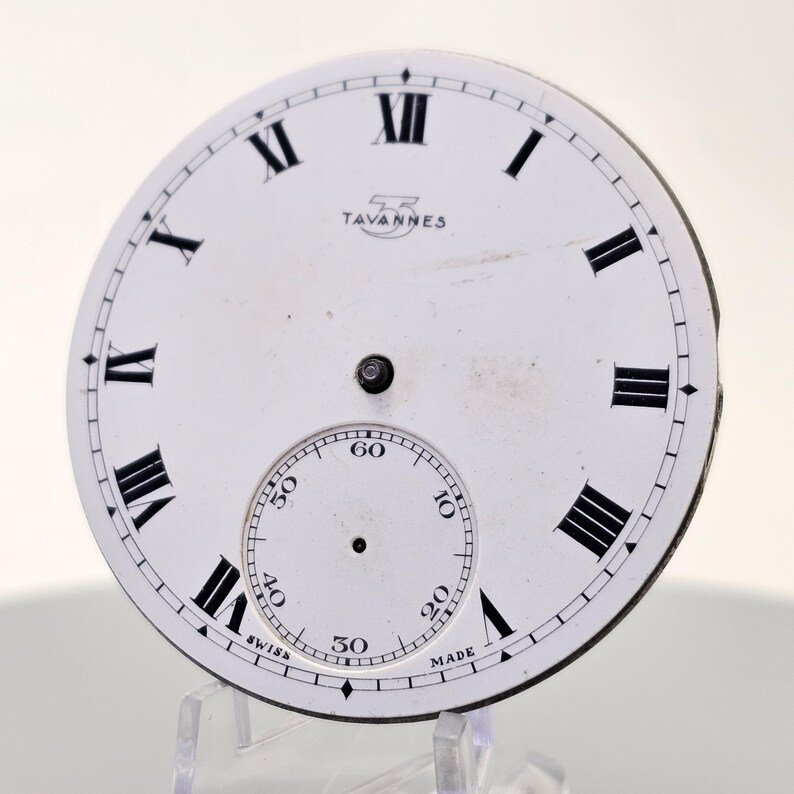 May include: A white watch face with black Roman numerals and a smaller seconds dial. The text "TAVANNES" is printed above the center. The words "SWISS MADE" are printed at the bottom of the seconds dial.