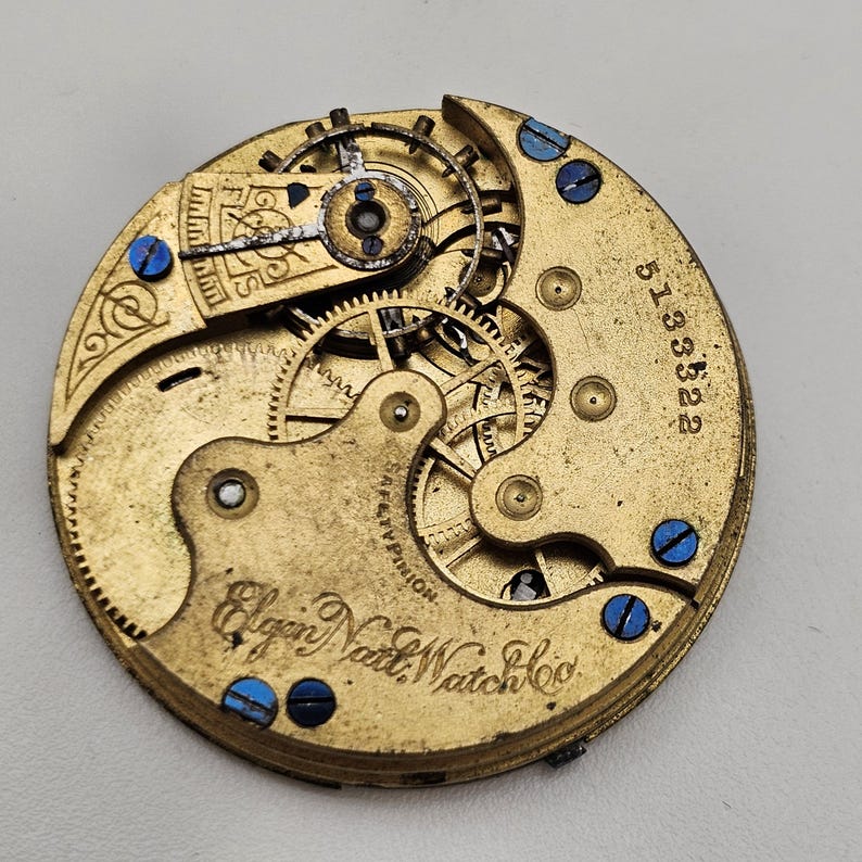 以下が含まれることがあります： ヴィンテージのElgin National Watch Co.の時計ムーブメントのクローズアップ。金色のメカニズムは、複雑な歯車、青いネジ、シリアル番号513322を備えています。20世紀初頭の時計製造の詳細な例です。