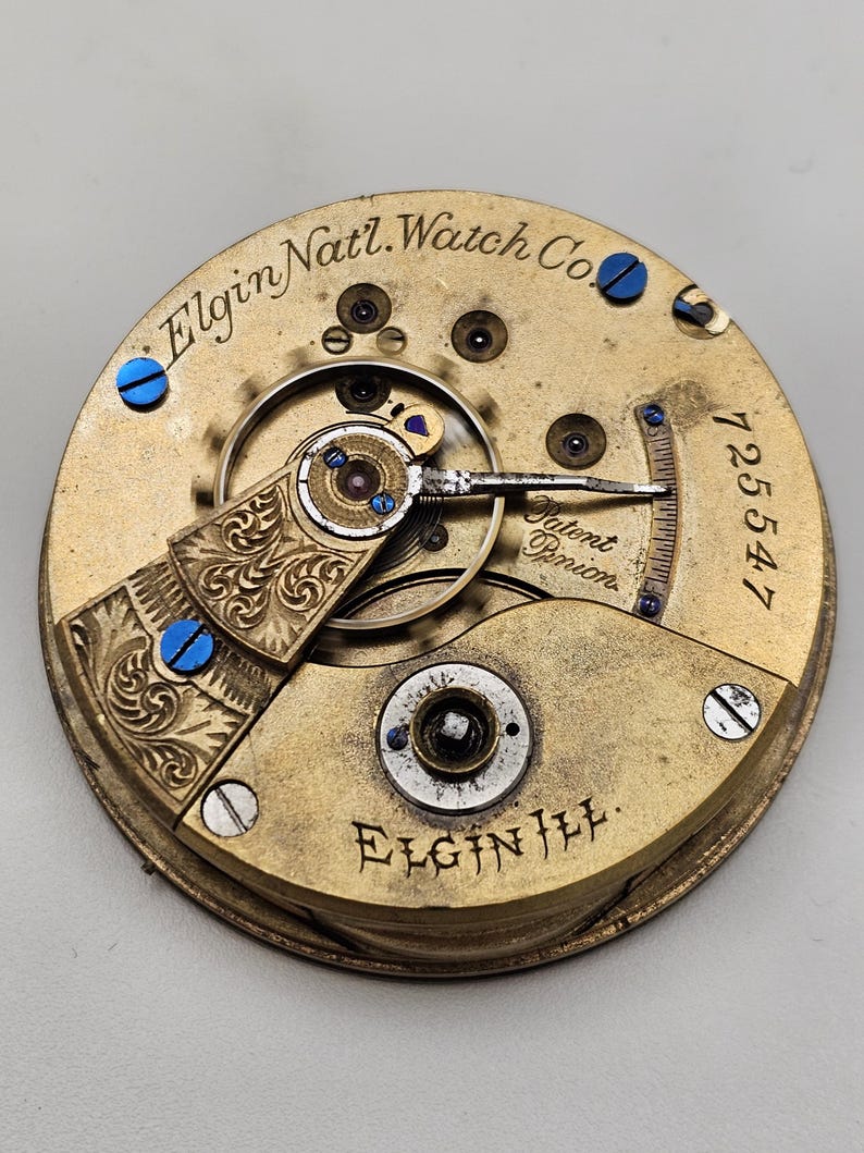 May include: Close-up of an Elgin Nat'l. Watch Co. pocket watch movement. The gold-toned mechanism features intricate details, including engraved patterns, blue screws, and the text "Elgin Nat'l. Watch Co." and "Elgin Ill.". The serial number "125547" is also visible.
