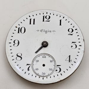 May include: A white pocket watch face with black numerals and hands. The word "Elgin" is printed in the center. A smaller dial is at the bottom, with numbers indicating seconds.