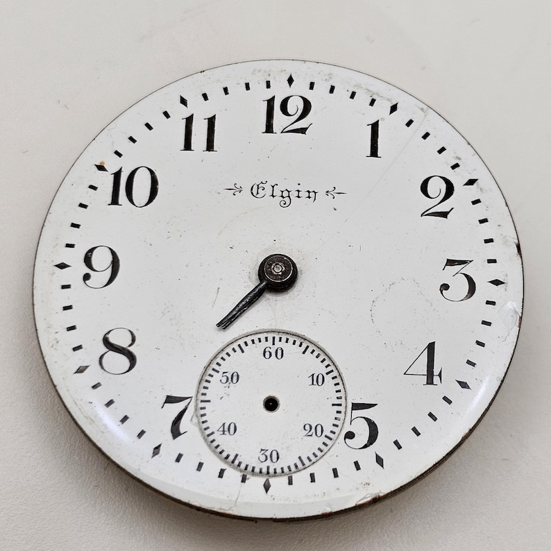 May include: A white pocket watch face with black numerals and hands. The word "Elgin" is printed in the center. A smaller dial is at the bottom, with numbers indicating seconds.