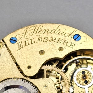May include: Close-up of a gold-toned pocket watch movement with the inscription "A Hendrick Ellesmere".