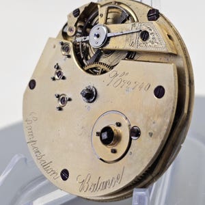 Może przedstawiać: Zbliżenie na mechanizm kieszonkowego zegarka w kolorze złotym z misternymi kołami zębatymi i wahadłem. Mechanizm jest opisany numerem "109940" oraz słowami "Compensation" i "Balance".