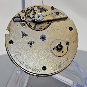 Może przedstawiać: Zbliżenie na mechanizm kieszonkowego zegarka w kolorze złotym z misternymi kołami zębatymi i wahadłem. Zegarek jest oznaczony jako "Compensation" i "18 Balance" oraz ma wygrawerowany numer "9672740" na płytce.