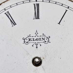 以下が含まれることがあります： 白い背景に、ヴィンテージの時計の文字盤をクローズアップ。黒いローマ数字が時間を表し、数字のXIIが目立っています。「ELGIN」の文字は装飾的なフォントです。下部に小さな穴があります。