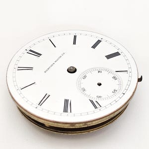 Puede incluir: Una esfera de reloj de bolsillo vintage con fondo blanco, n&uacute;meros romanos negros y un peque&ntilde;o dial de segundos. La esfera del reloj tiene el texto "HAMPDEN WATCH CO" y un borde met&aacute;lico.