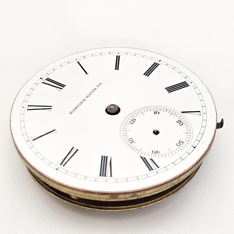 Puede incluir: Una esfera de reloj de bolsillo vintage con fondo blanco, n&uacute;meros romanos negros y un peque&ntilde;o dial de segundos. La esfera del reloj tiene el texto "HAMPDEN WATCH CO" y un borde met&aacute;lico.