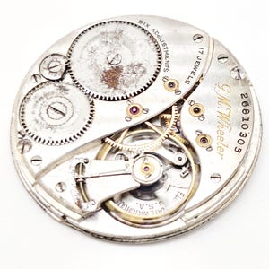 May include: Close-up of a vintage watch movement with intricate gears, wheels, and jewels. The watch mechanism is silver-toned with gold accents and the text "G.M. Wheeler" and "17 Jewels". The watch also has the text "Six Adjustments" and "U.S.A."