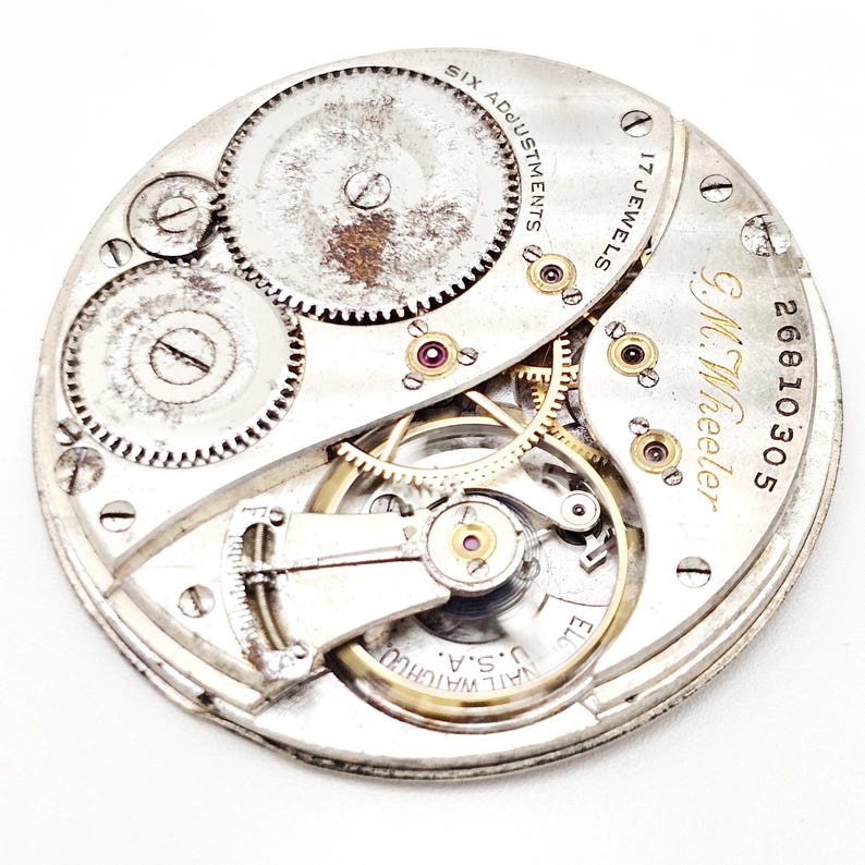 May include: Close-up of a vintage watch movement with intricate gears, wheels, and jewels. The watch mechanism is silver-toned with gold accents and the text "G.M. Wheeler" and "17 Jewels". The watch also has the text "Six Adjustments" and "U.S.A."