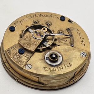 May include: Close-up of an antique Elgin Nat'l. Watch Co. pocket watch movement. The gold-toned mechanism features intricate gears, engraved details, and the text "Patent Pinion". The serial number 725547 is visible.