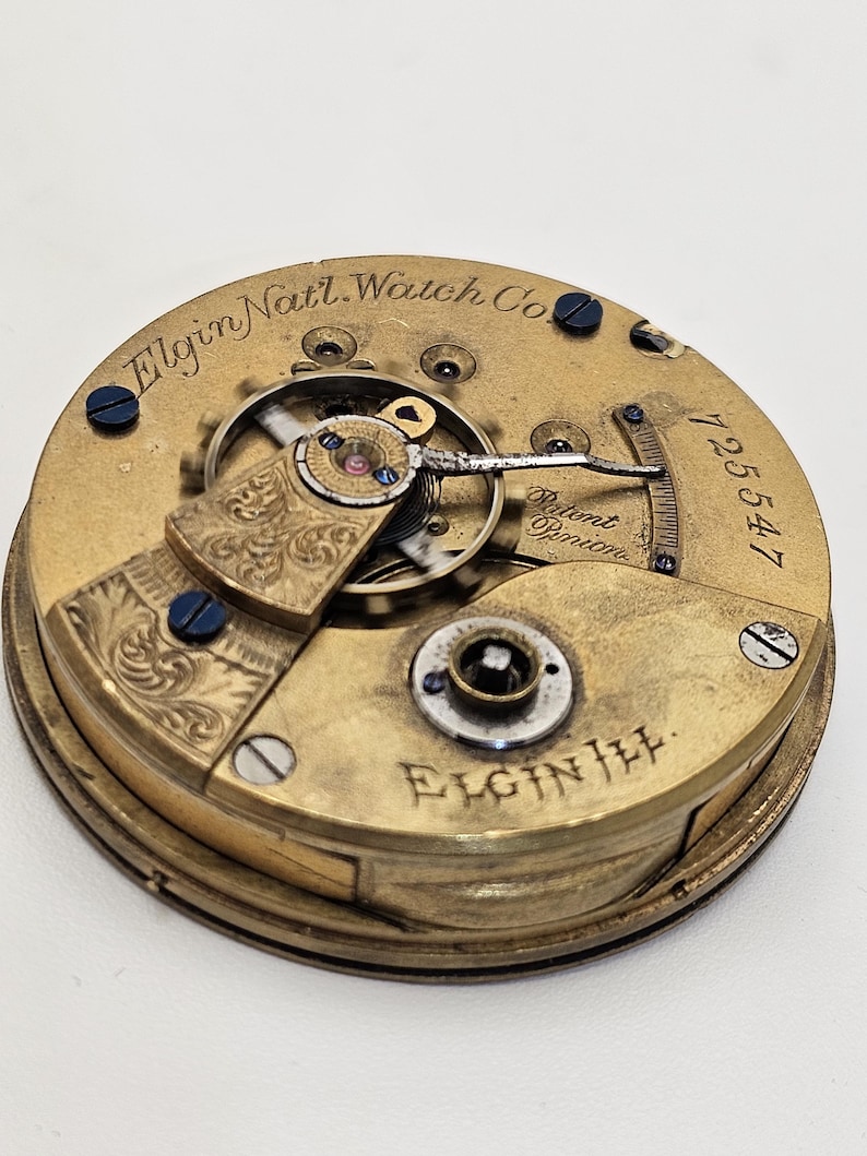 May include: Close-up of an antique Elgin Nat'l. Watch Co. pocket watch movement. The gold-toned mechanism features intricate gears, engraved details, and the text "Patent Pinion". The serial number 725547 is visible.